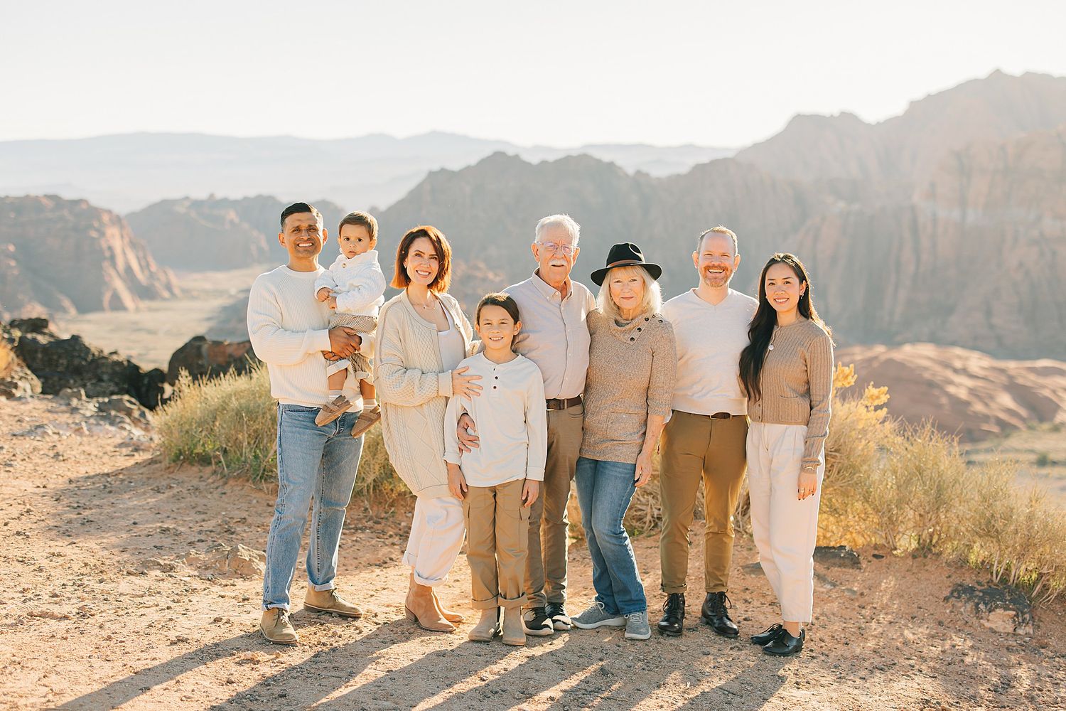 Extended family of eight smiles together in a desert setting with mountain backdrop in soft natural light.