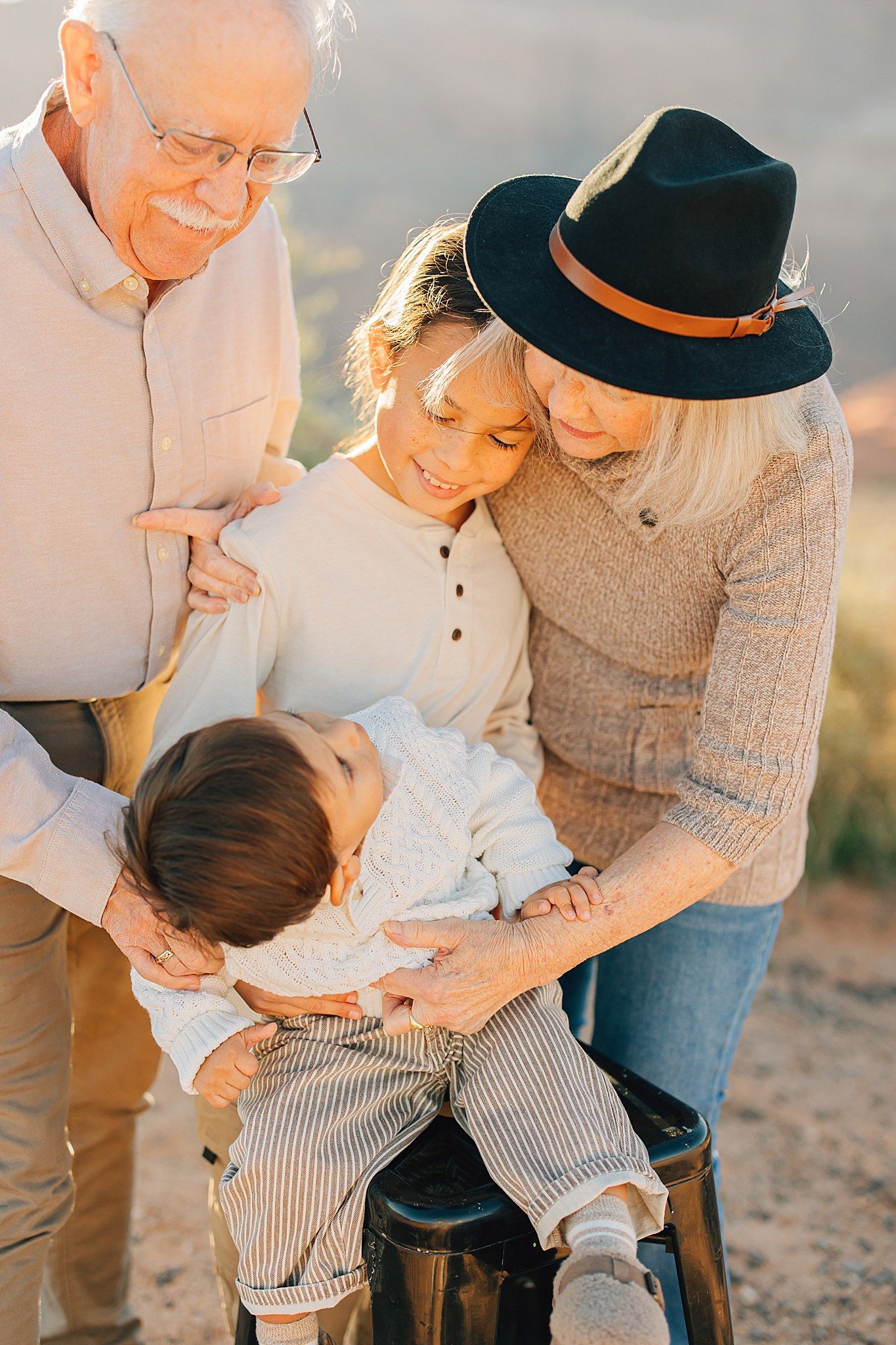 Grandparents and grandchildren share a tender moment outdoors in warm golden light, gathered around a toddler on a stool.