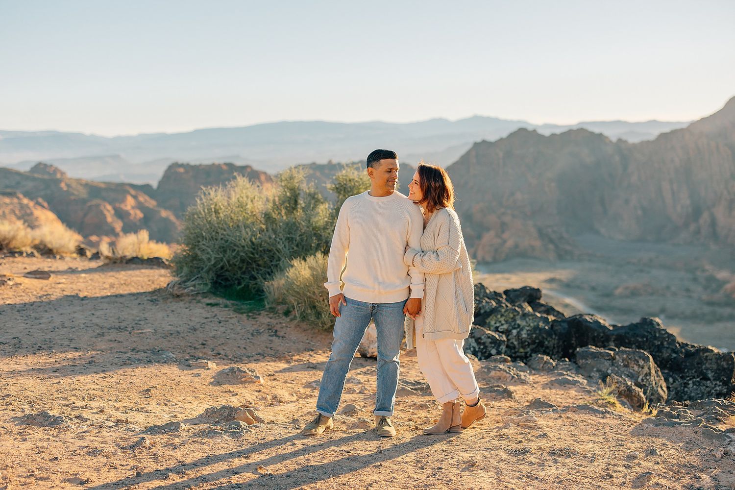 Couple holding hands and gazing at each other in a golden desert landscape with mountains in the background.