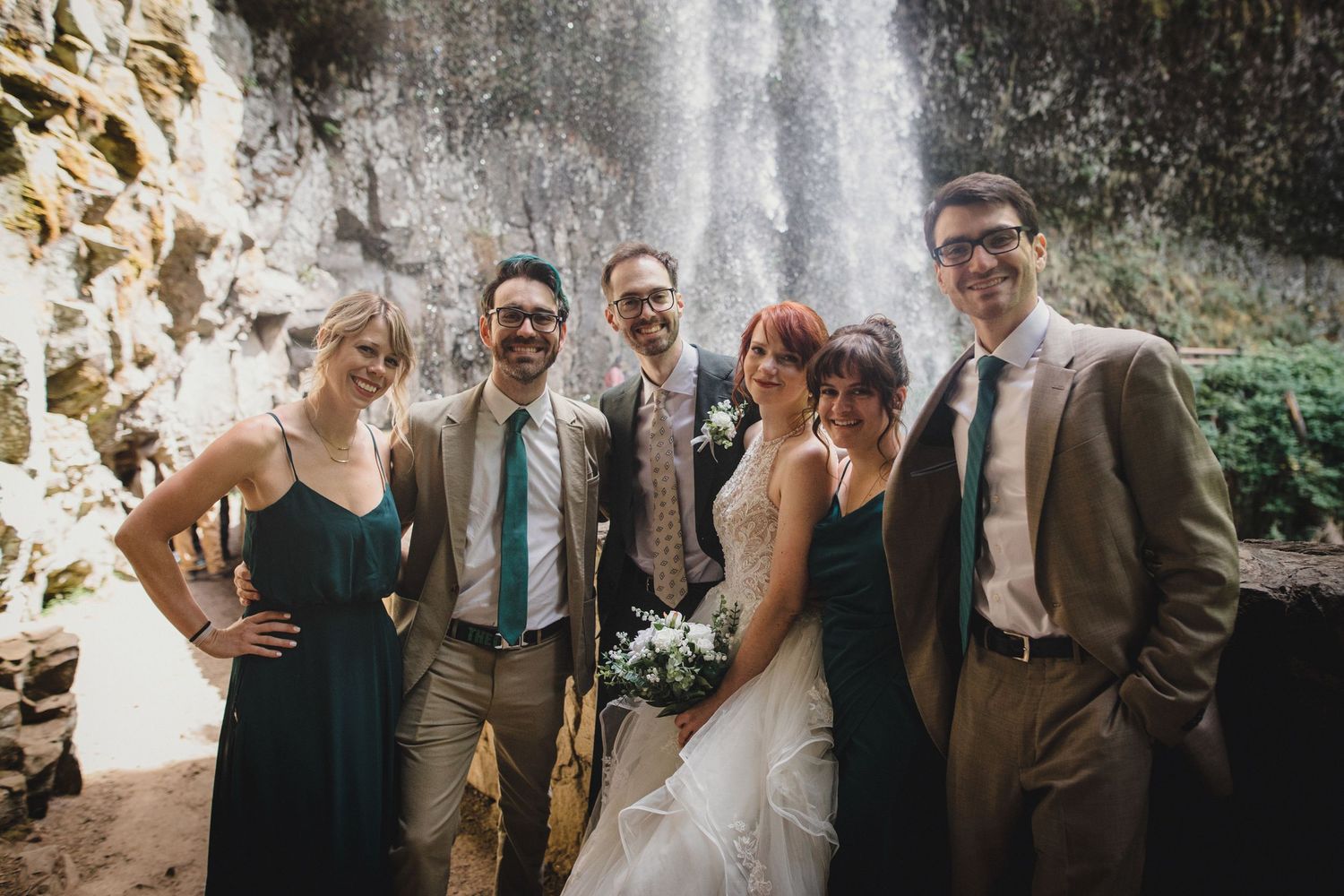Wedding party of six smiling in front of a waterfall, bridesmaids in teal dresses, groomsmen in tan suits.