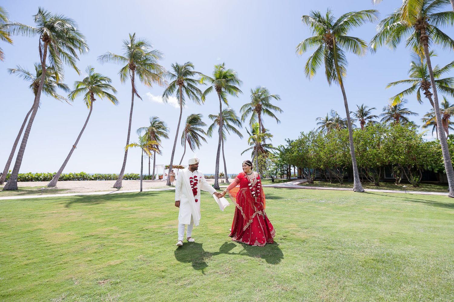 A bride and groom dressed in traditional Indian wedding attire stand in a tropical garden setting.