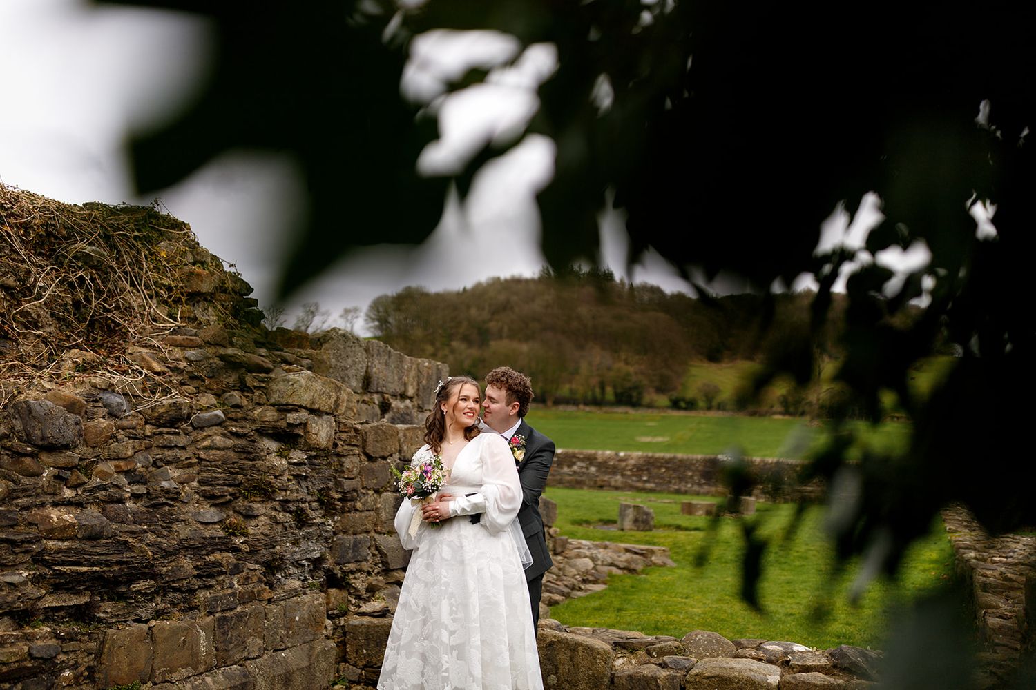 A bride and groom embrace in a picturesque outdoor setting with a stone wall and lush greenery in the background.