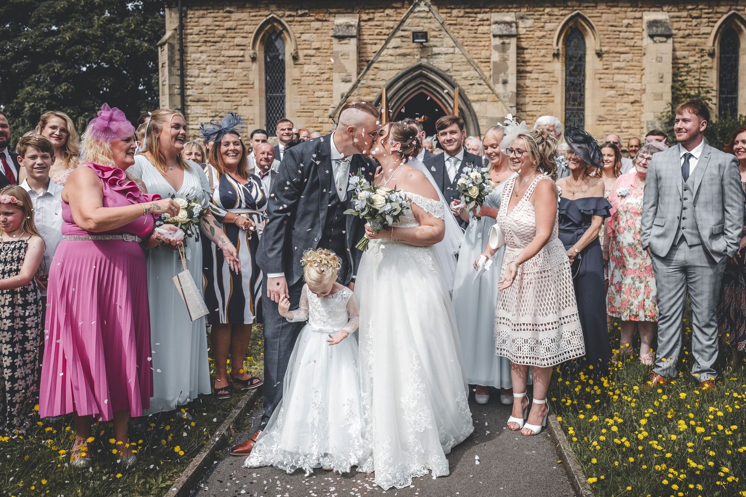Wedding guests shower confetti on newlyweds outside a stone church with gothic architecture.