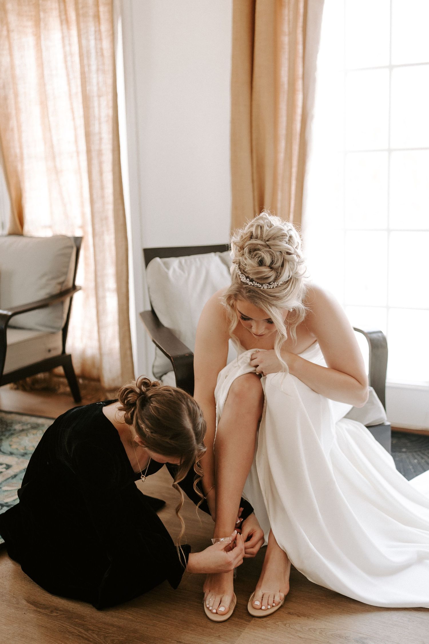 Bridesmaid kneeling to help bride put on sandals before winter wedding ceremony.