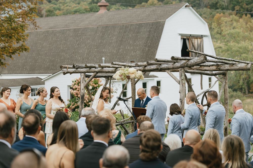 A rustic outdoor wedding ceremony takes place in front of a white barn under a wooden arbor on a sunny autumn day.