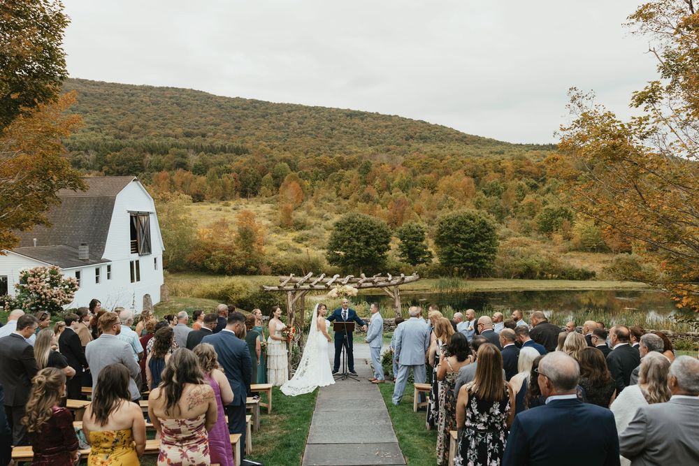 Panoramic view of an outdoor wedding ceremony in a scenic mountain setting with guests seated on white chairs.