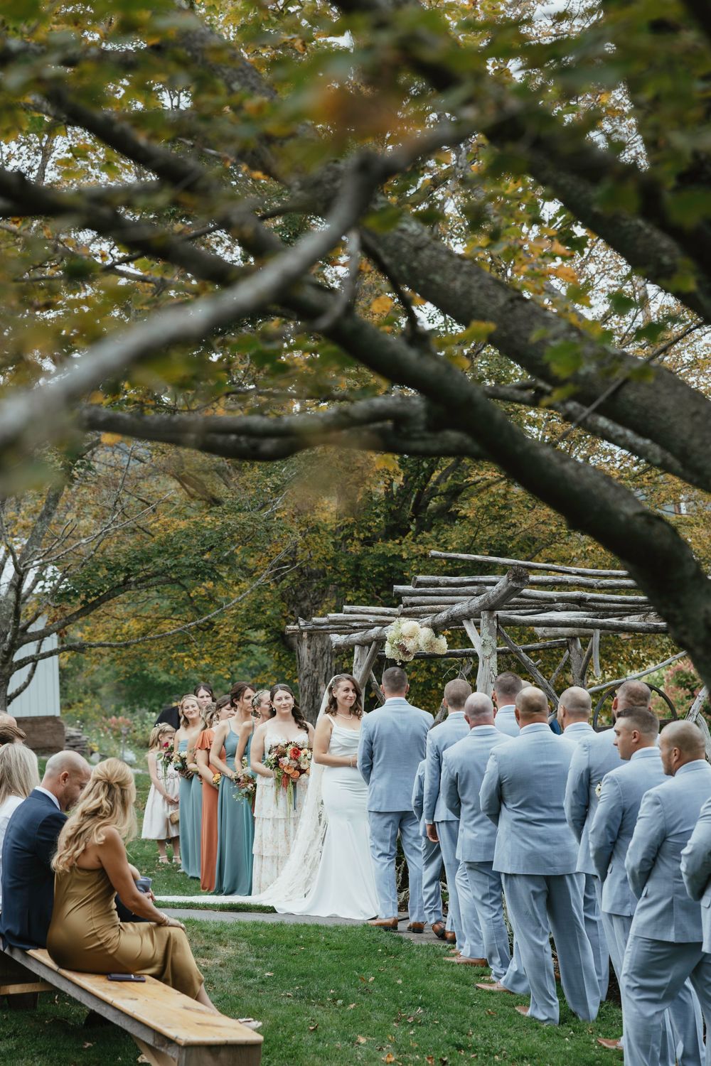 Wedding ceremony guests stand in rows outdoors under autumn trees during a fall celebration.
