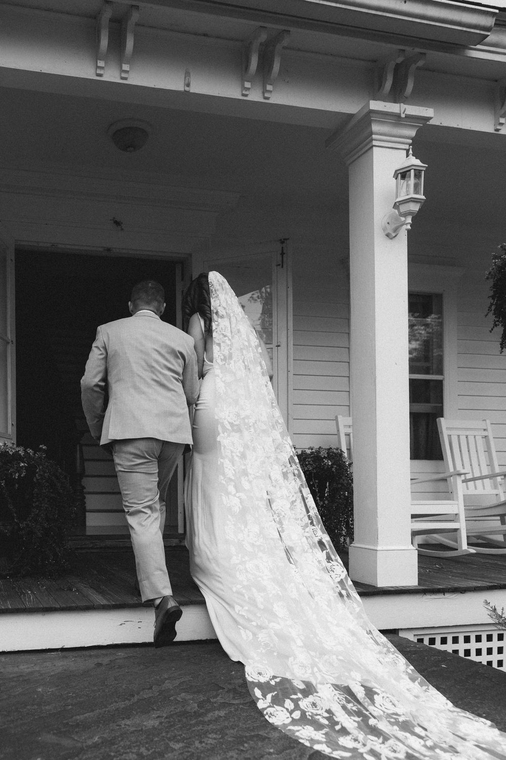 Black and white photo of a flowing white wedding dress train following across a porch with rocking chairs.