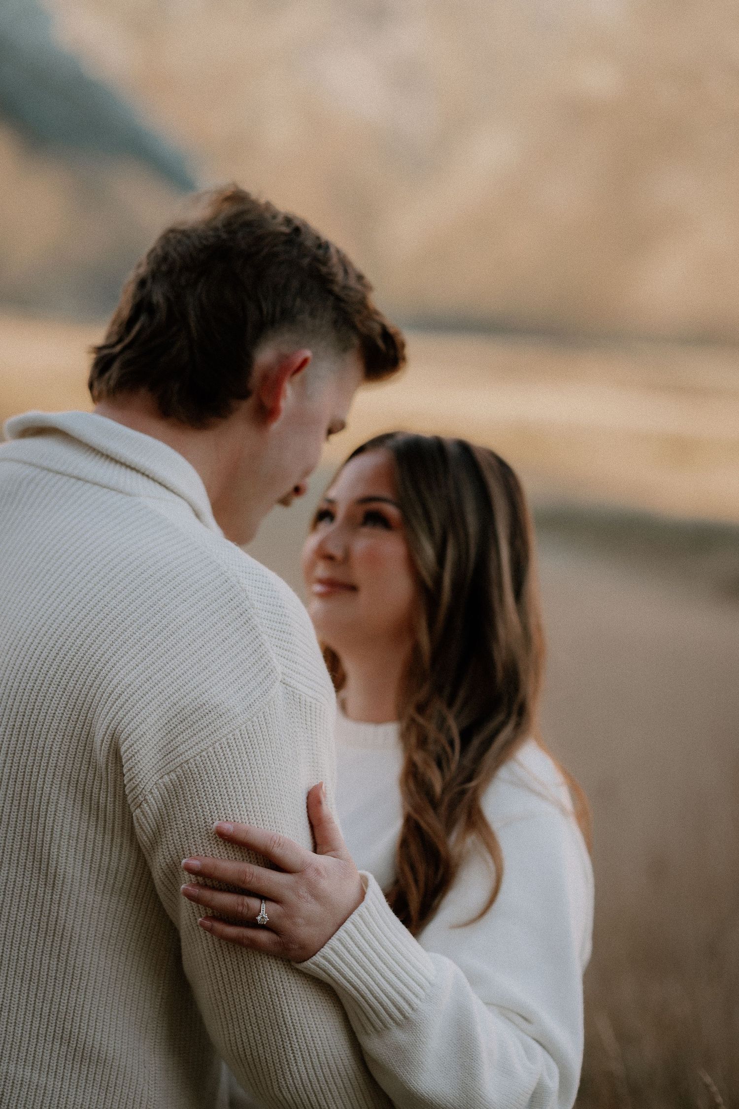 A couple embraces in a golden wheat field at sunset with mountains in the background creating a romantic silhouette.