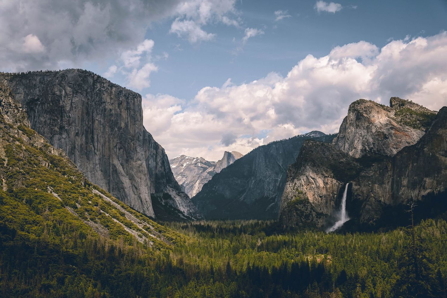Panoramic view of Yosemite Valley with El Capitan, Bridalveil Fall, and lush forest under dramatic cloudy skies.