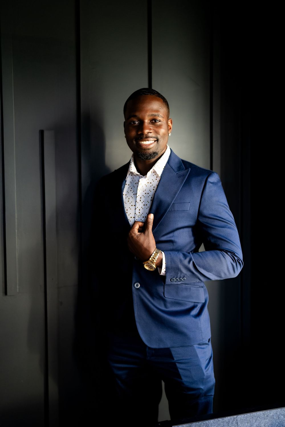 Smiling Black man in blue suit and gold watch leaning against dark wall in professional portrait photo.