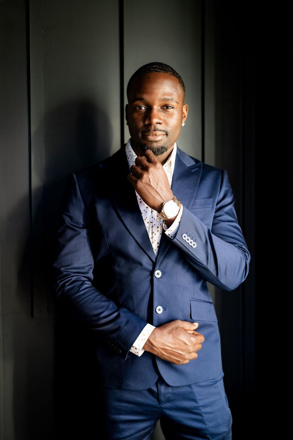Professional Black man in navy blue suit posing confidently against dark wall, wearing a watch and patterned shirt.