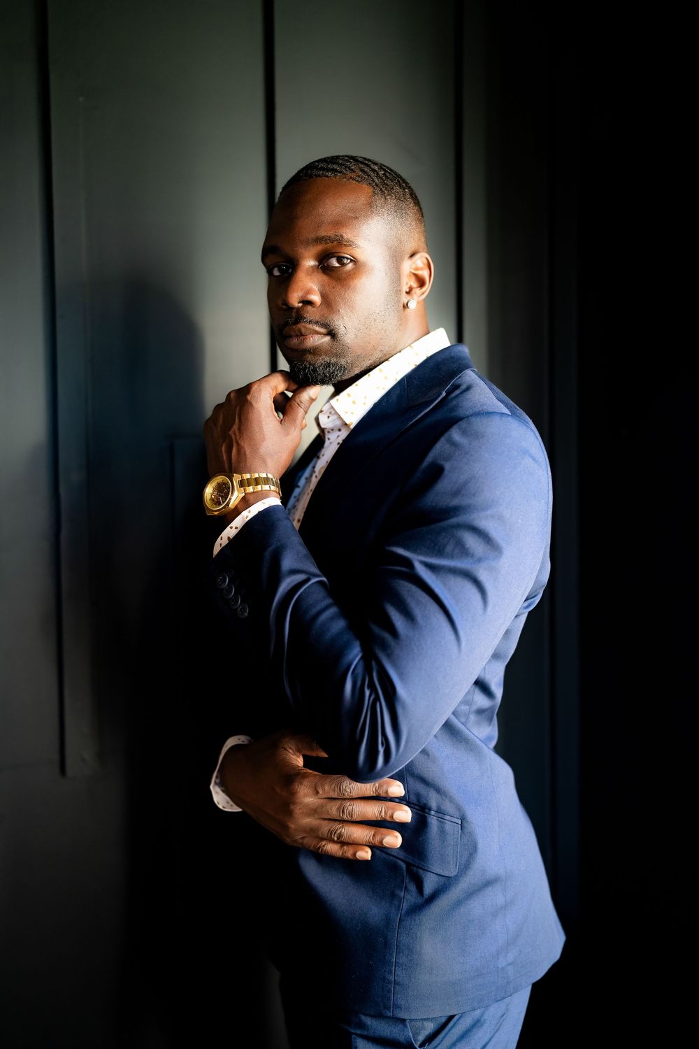 A well-dressed Black man in a blue suit and gold watch poses confidently against a dark wall.