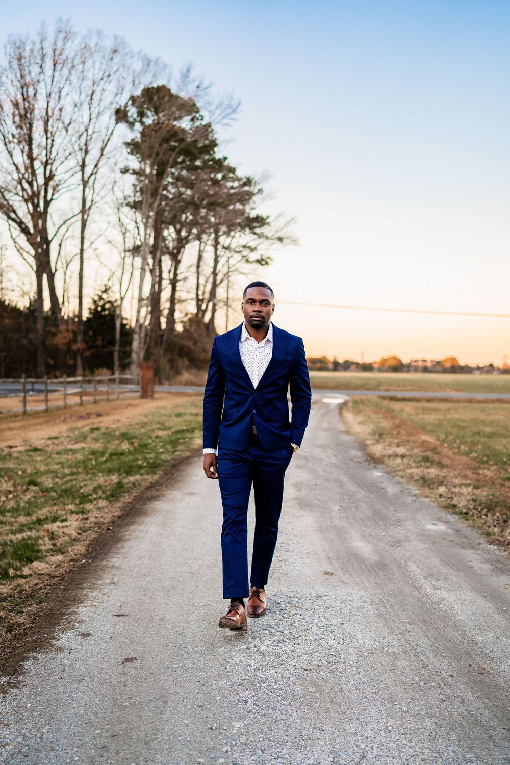 A well-dressed Black man in a navy blue suit walks confidently down a rural gravel road at sunset.