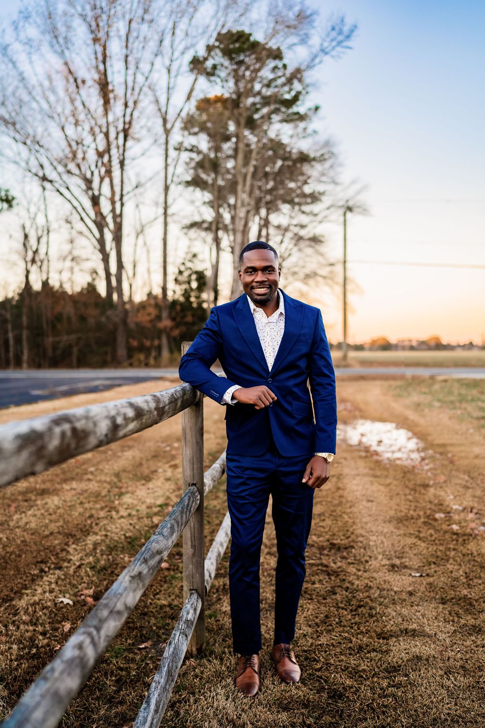 Smiling man in navy blue suit leaning on wooden fence outdoors at golden hour.