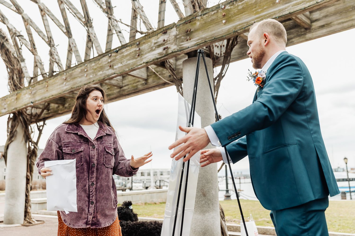 Surprised woman reacts with open arms to a man in a teal suit under a wooden pergola outdoors.