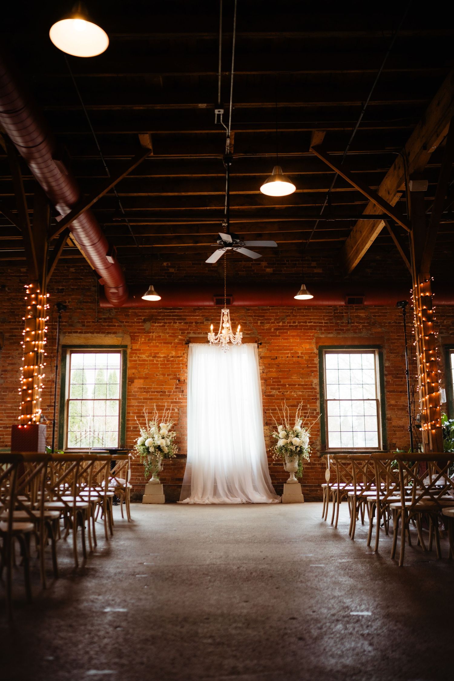 Elegant rustic wedding ceremony setup with white draped arch, floral arrangements, and string lights in brick venue.
