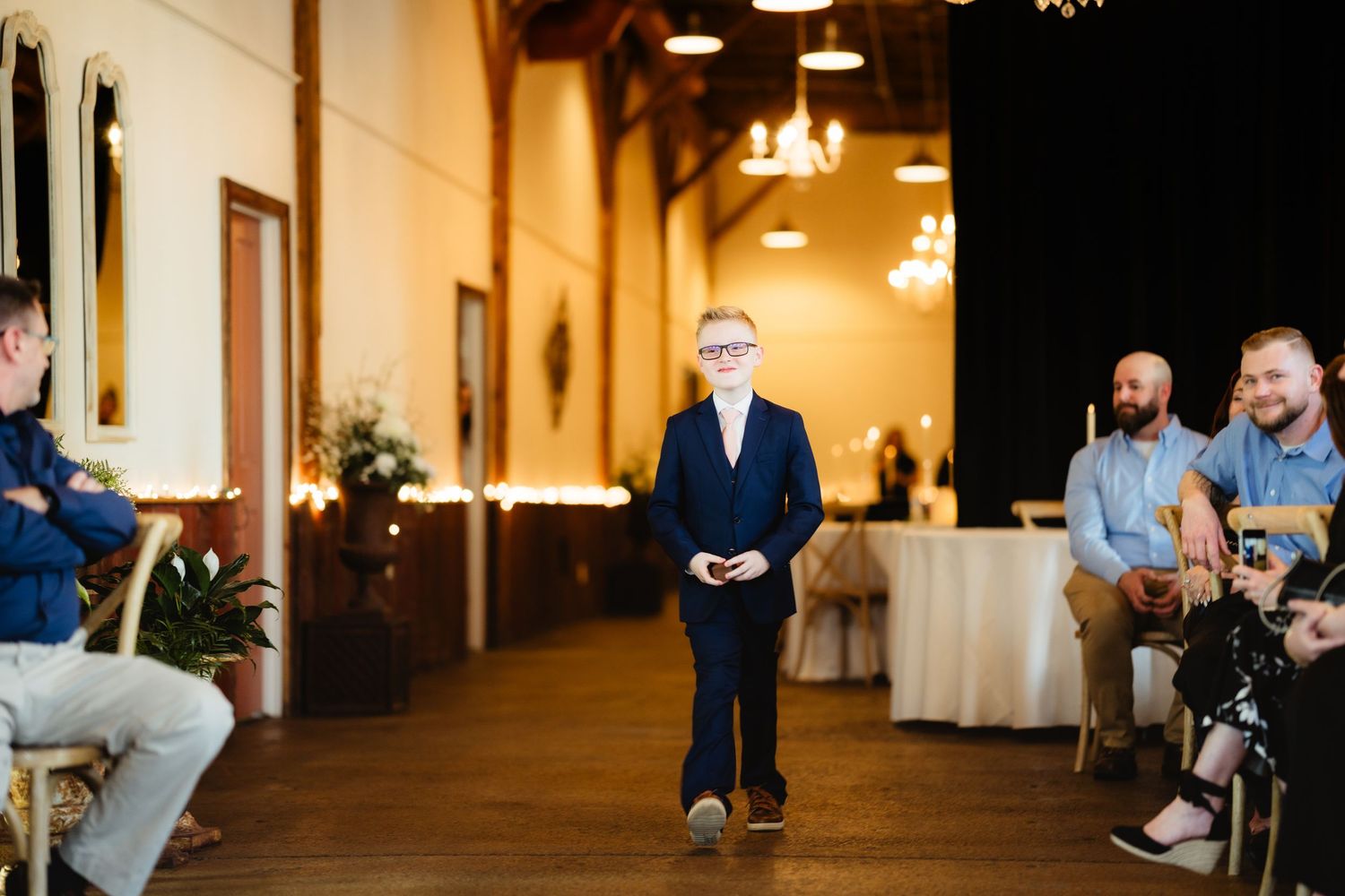 Young ring bearer in navy suit walks down wedding aisle while guests smile and applaud in elegant venue.