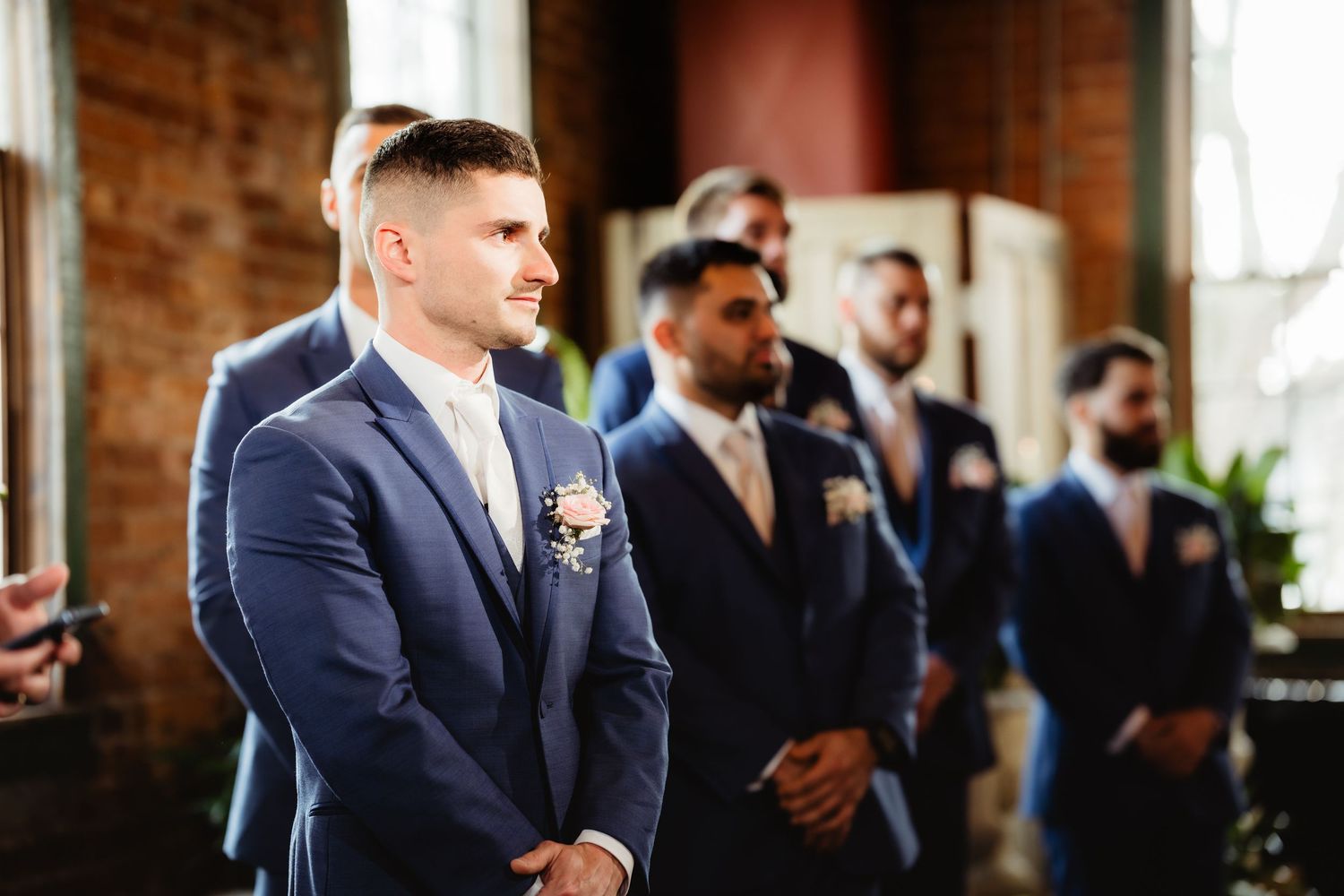 Groom and groomsmen in navy blue suits with boutonnieres standing at wedding altar in brick venue.