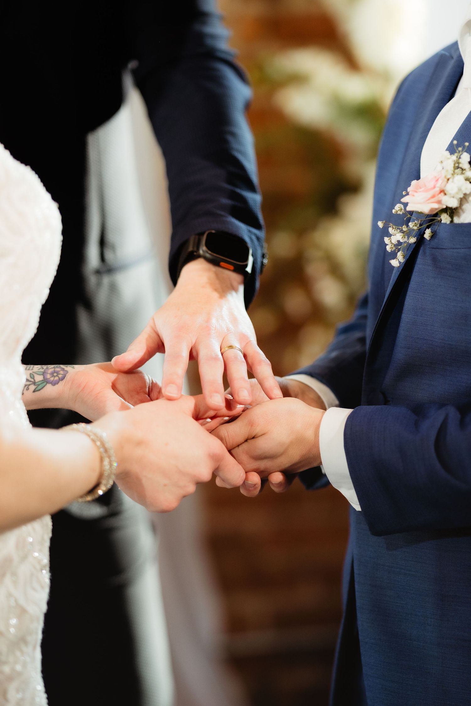 Bride and groom exchanging wedding rings during ceremony, close-up of hands with tattoo and boutonniere visible.