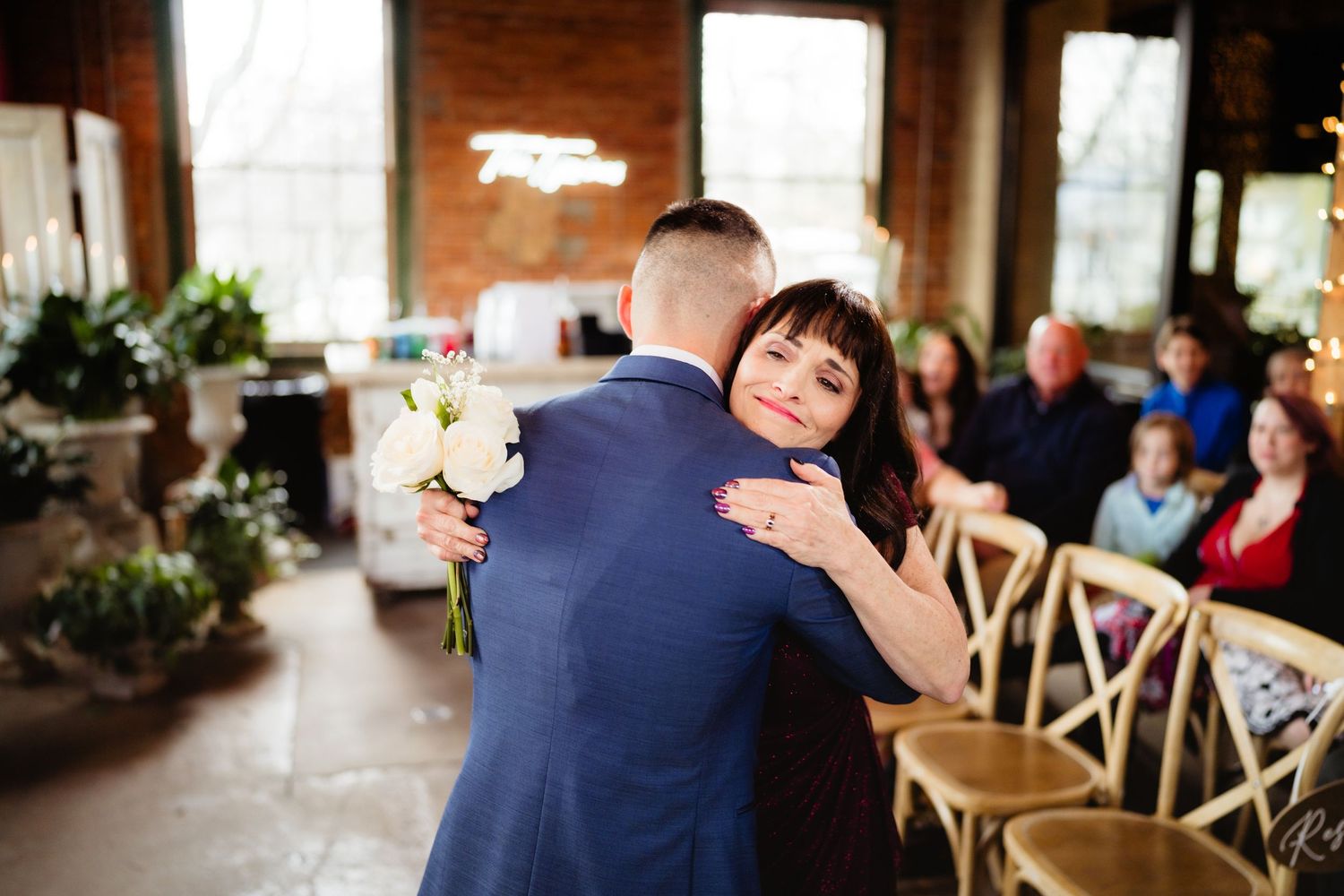 Bride and groom share a heartfelt embrace during indoor wedding ceremony with guests seated on wooden chairs.