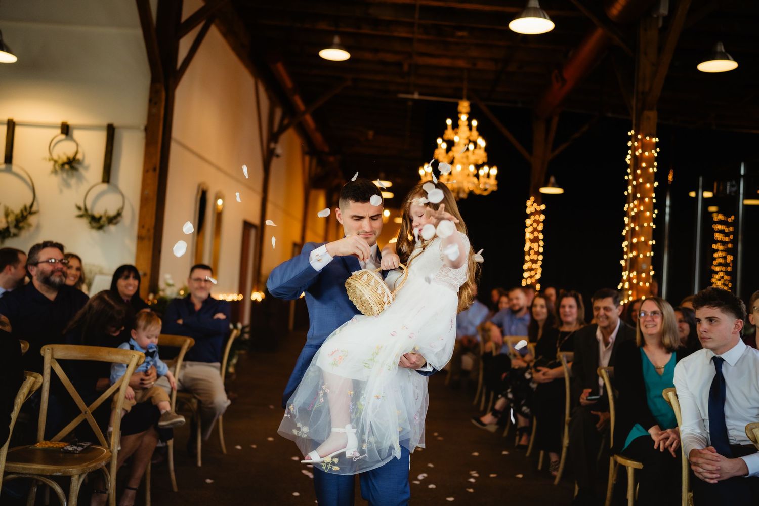 Groom lifts flower girl at wedding ceremony in rustic barn venue with string lights and seated guests.