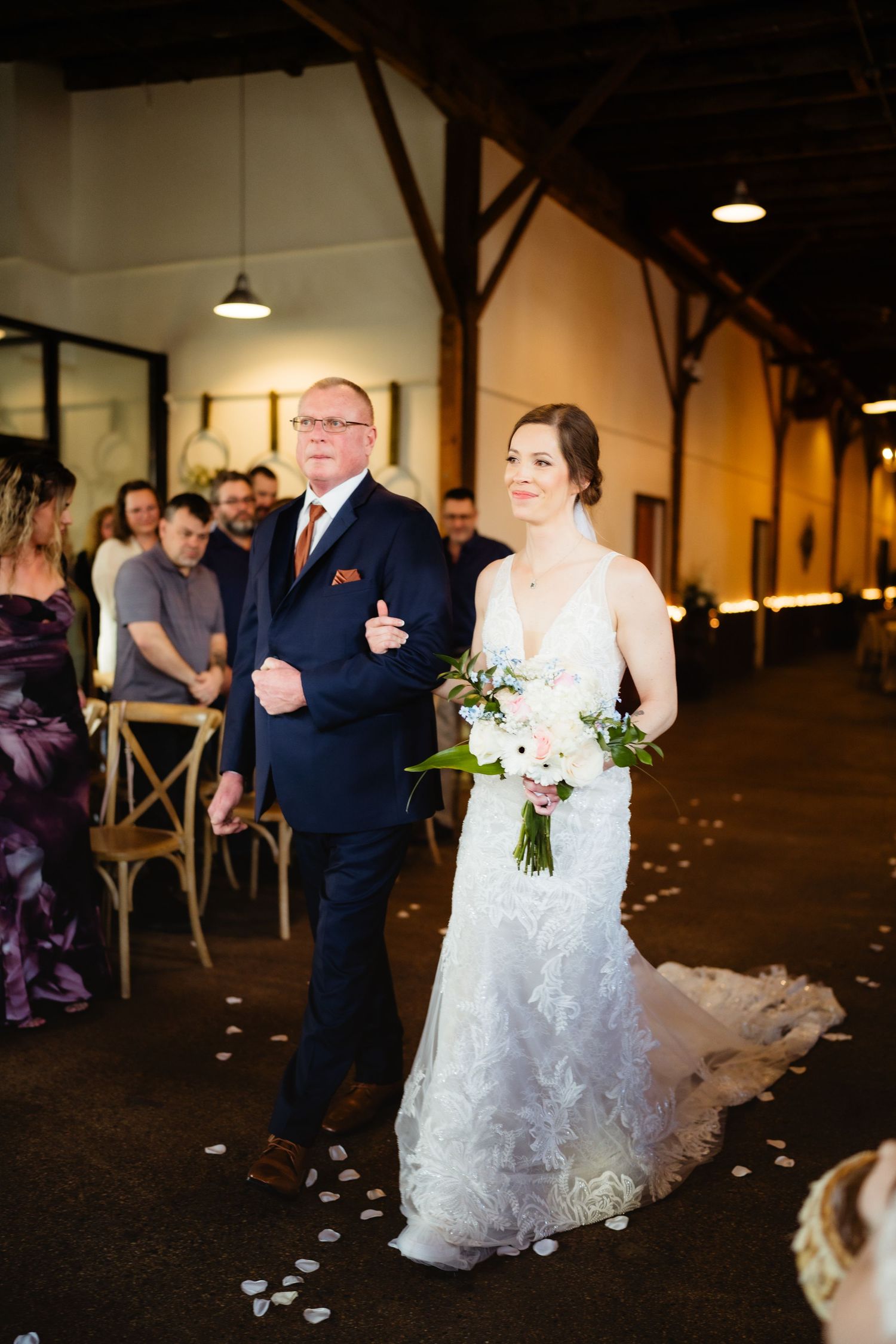 Bride in white lace gown walks down aisle with escort holding pink bouquet at indoor wedding ceremony.