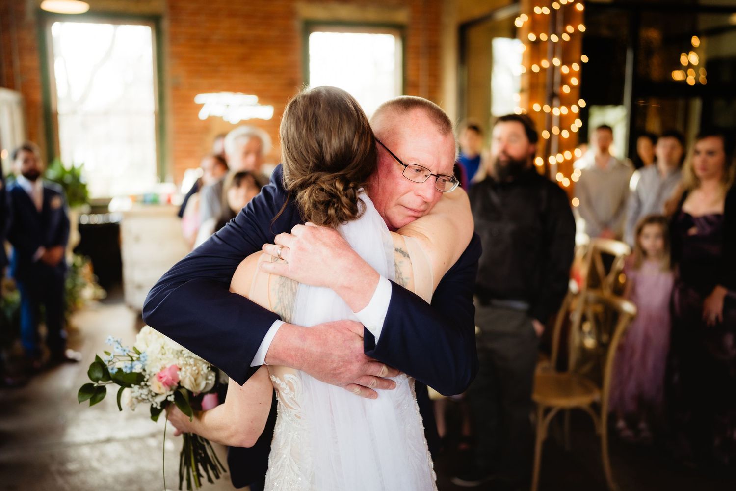 Emotional father-daughter wedding hug at indoor ceremony with string lights and guests in background.