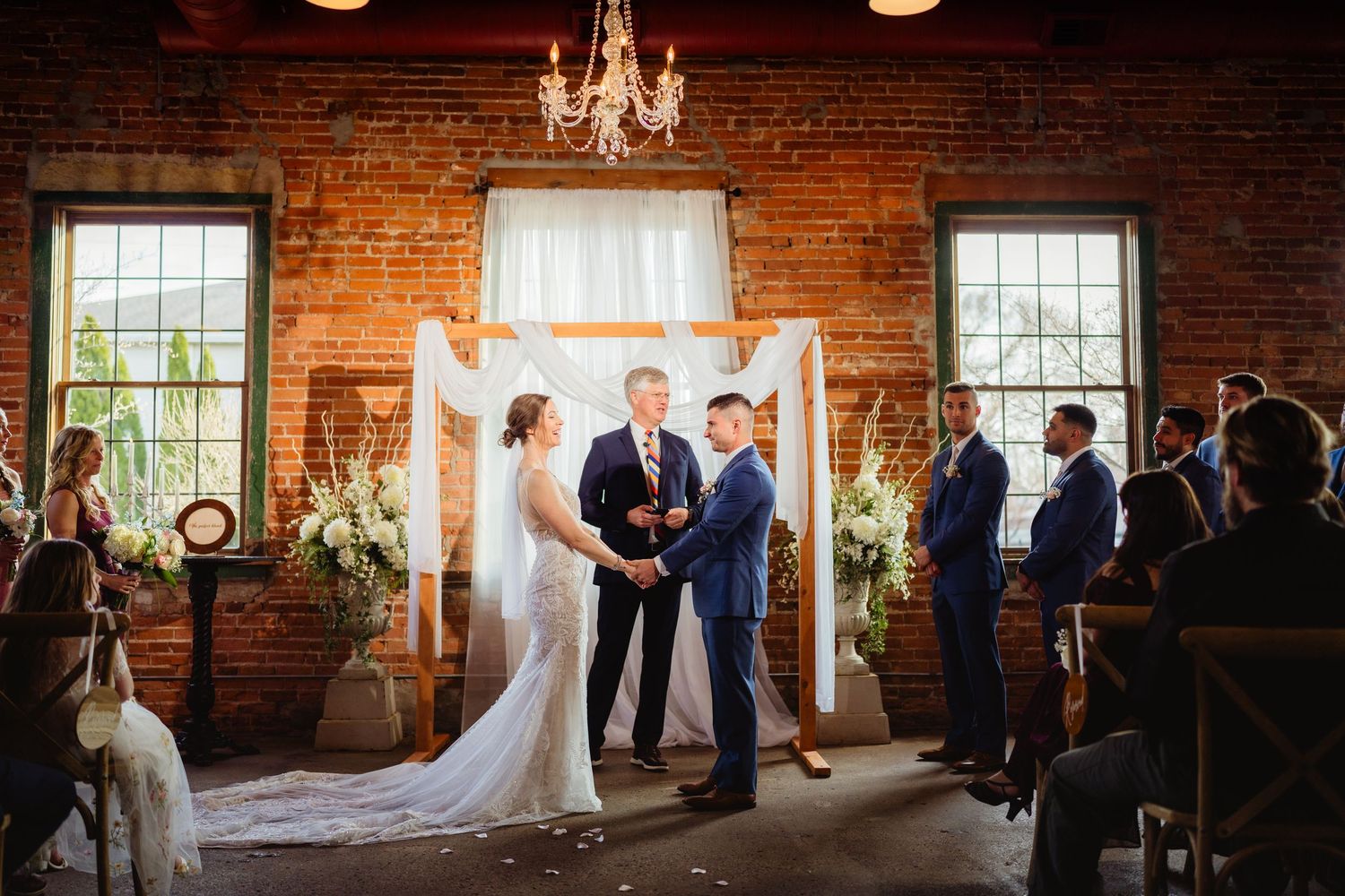 Couple exchanges vows at rustic brick wall wedding ceremony with white floral arch and chandelier.