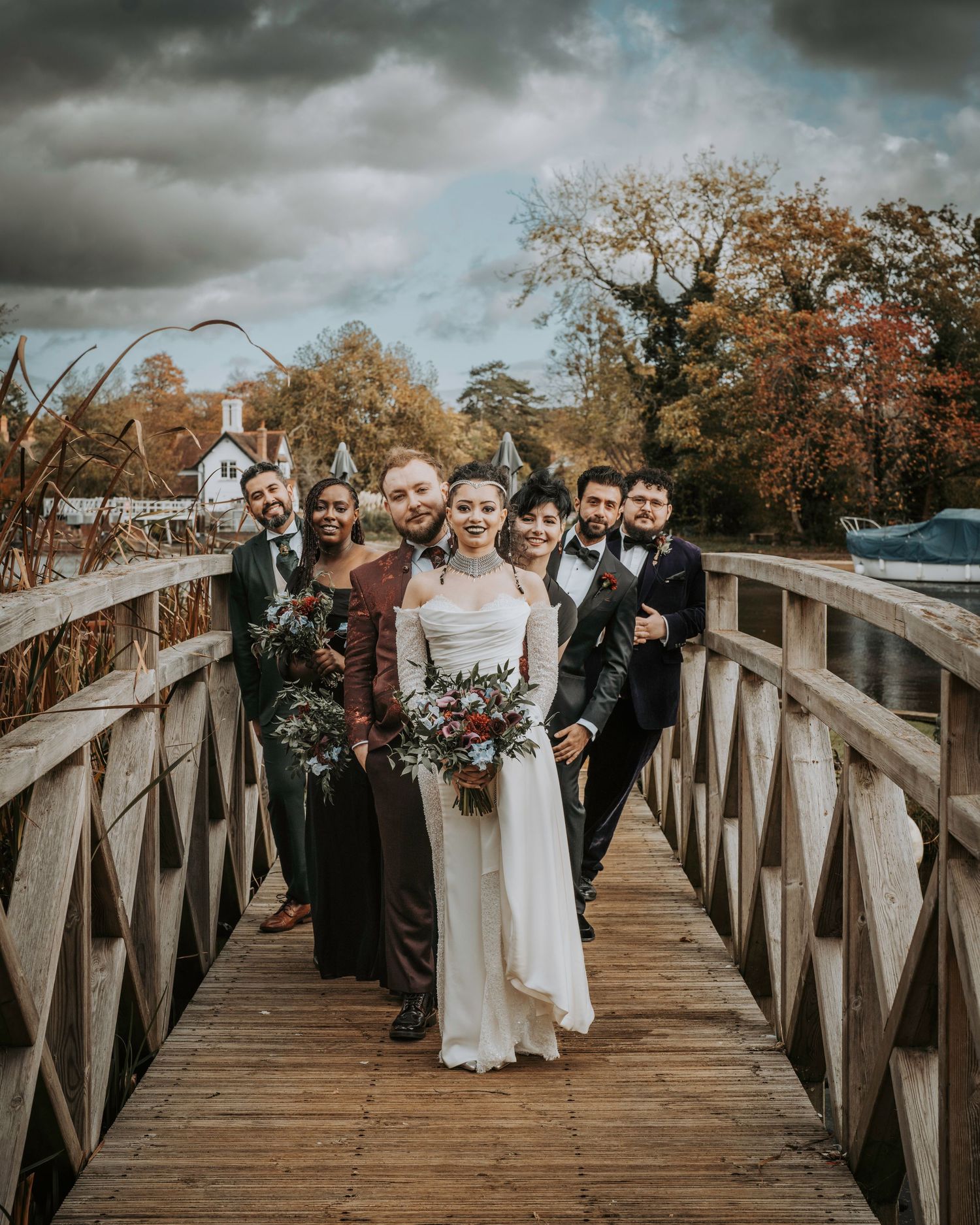Wedding party poses on wooden bridge with autumn foliage backdrop, bride in white gown holding red bouquet.