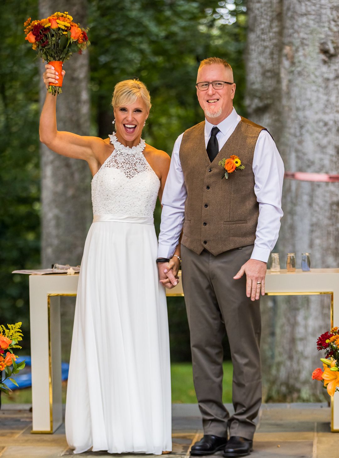 A couple in wedding attire celebrates their outdoor ceremony with joyful poses in front of tall trees.
