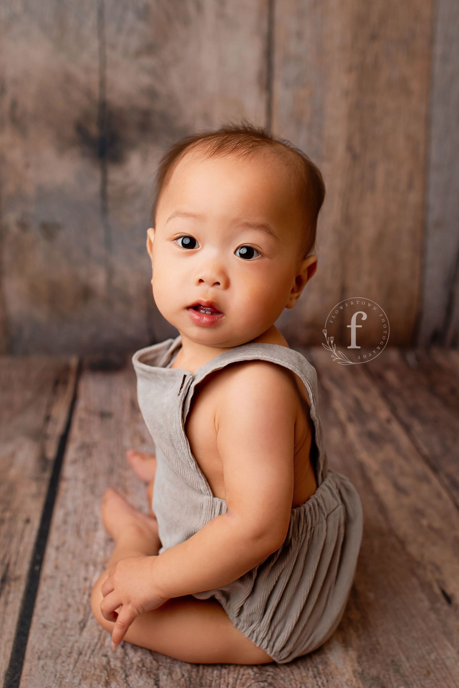 Baby boy in gray romper sitting on rustic wood floor looking over shoulder during sitter session at Flowertown Photography Summerville SC