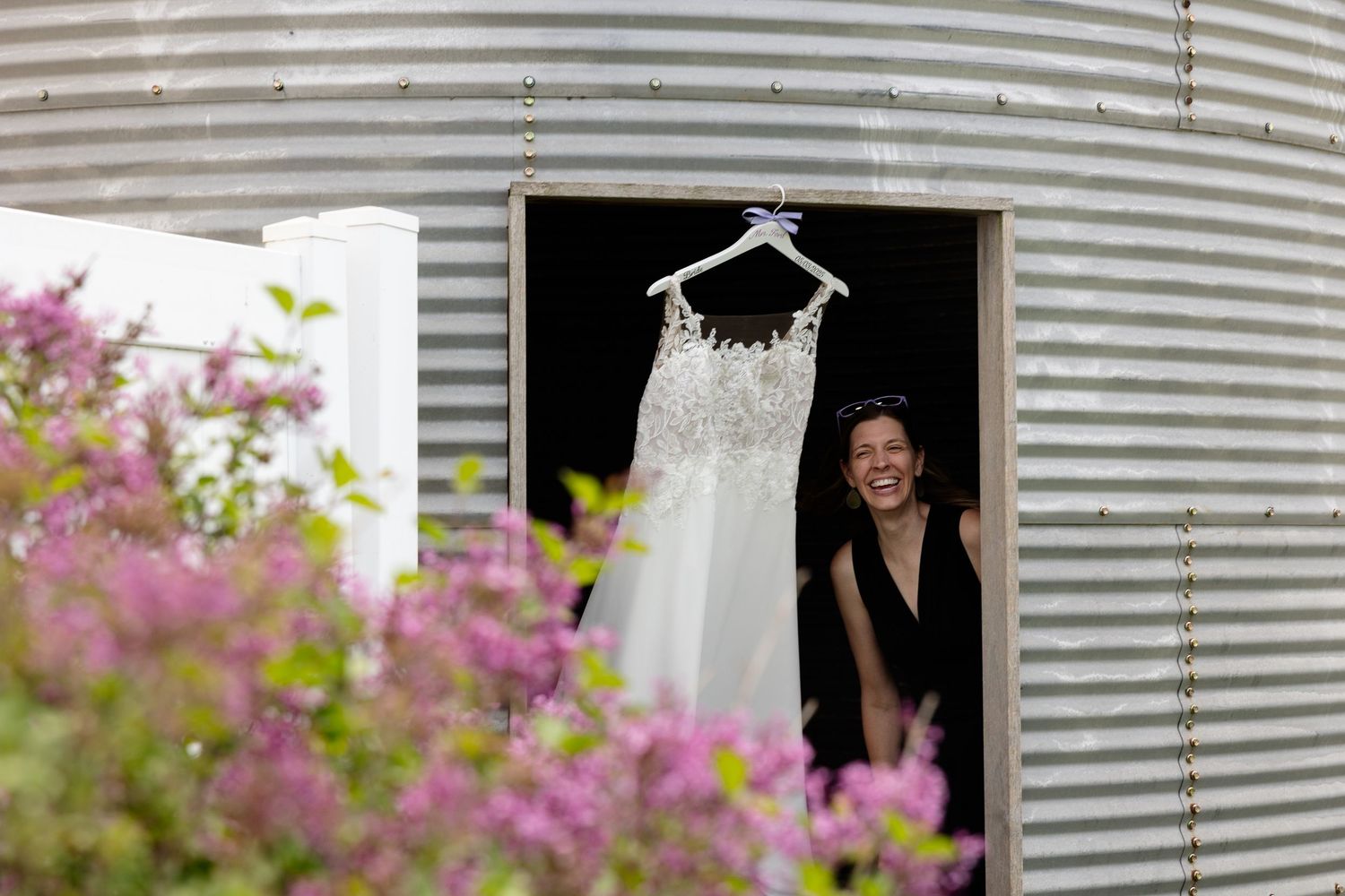 A white wedding dress hangs in a doorway of a grain silo with pink flowers in the foreground.