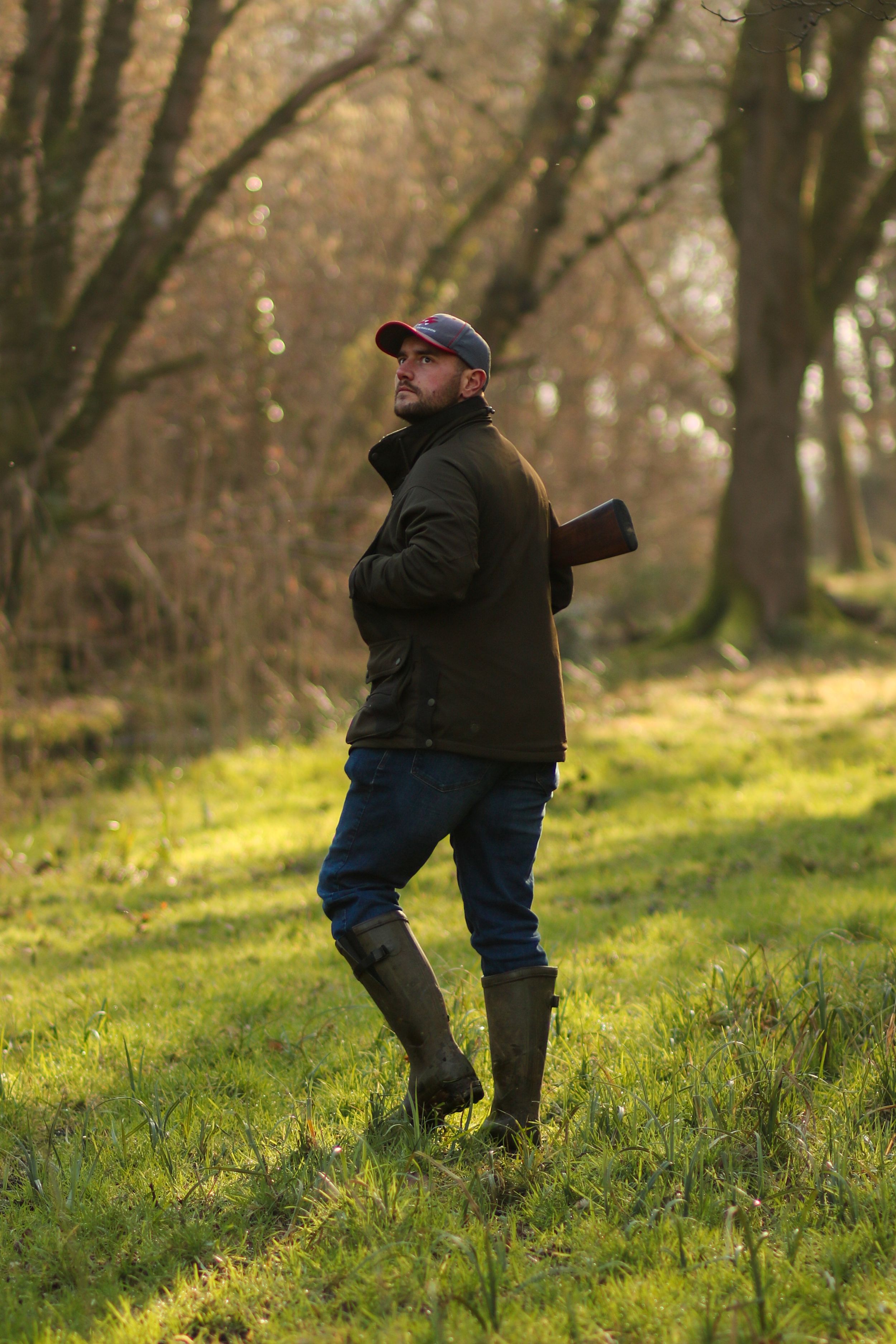 A male hunter carrying a shotgun walking through a sunlit woodland wearing wellington boots and a cap.