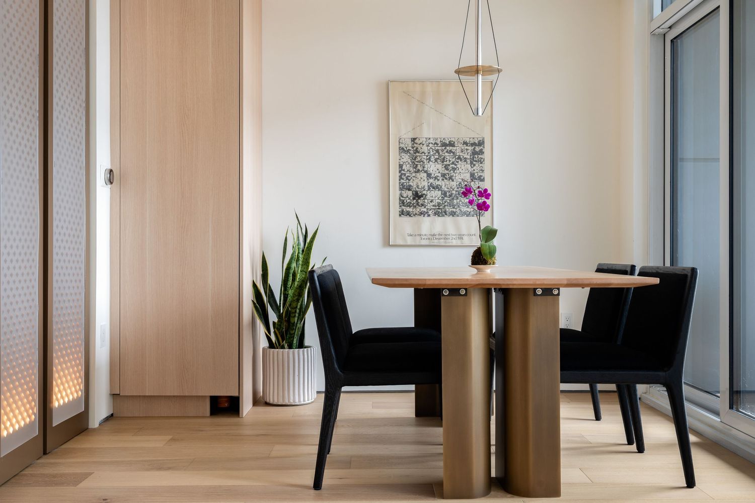 Minimalist dining area featuring round wooden table, black upholstered chairs, orchid bloom, abstract artwork, and geometric pendant light.