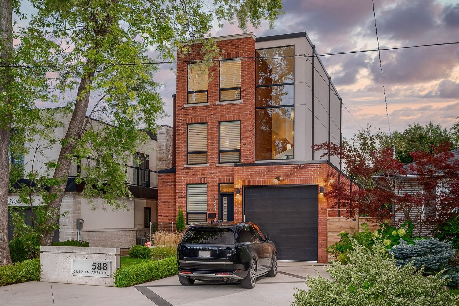 Modern three-story brick and glass home with attached garage and lush trees at dusk.