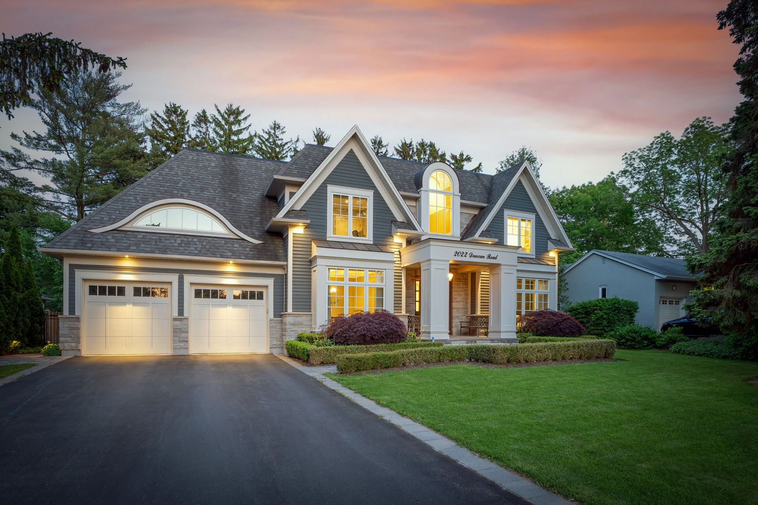 Elegant suburban home with gray roof, lit windows, manicured lawn, and lush trees at twilight sunset.