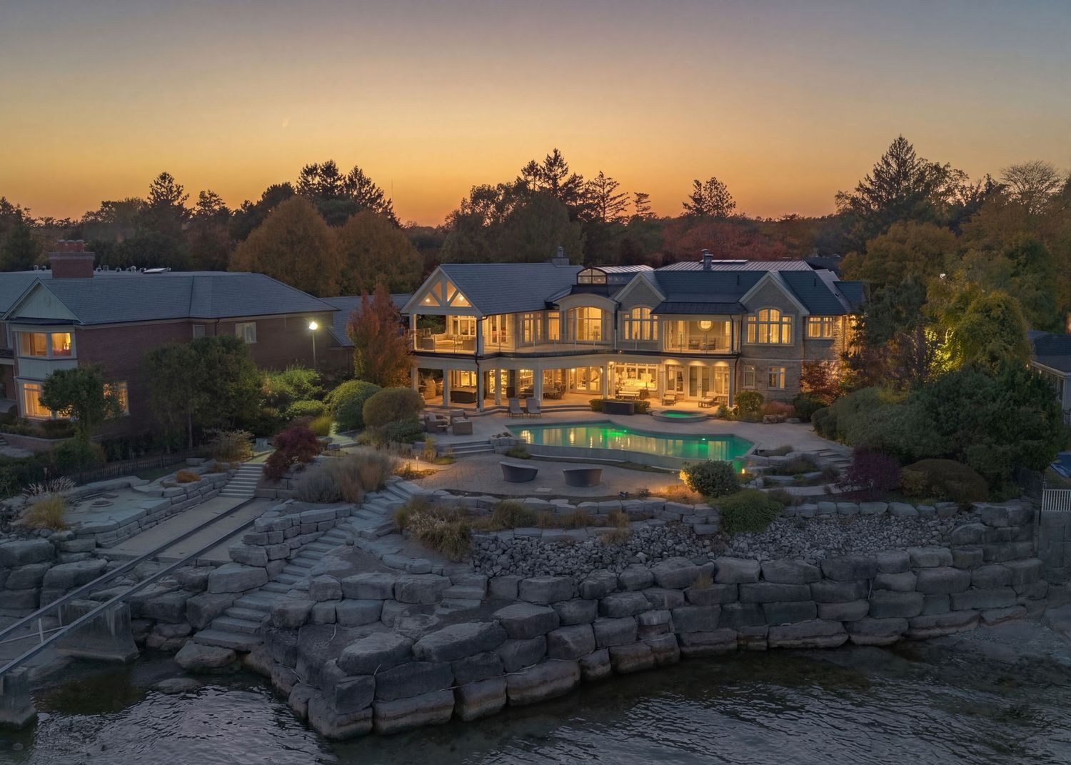 Luxurious waterfront mansion with infinity pool overlooking rocky shoreline at sunset with warm glowing lights.