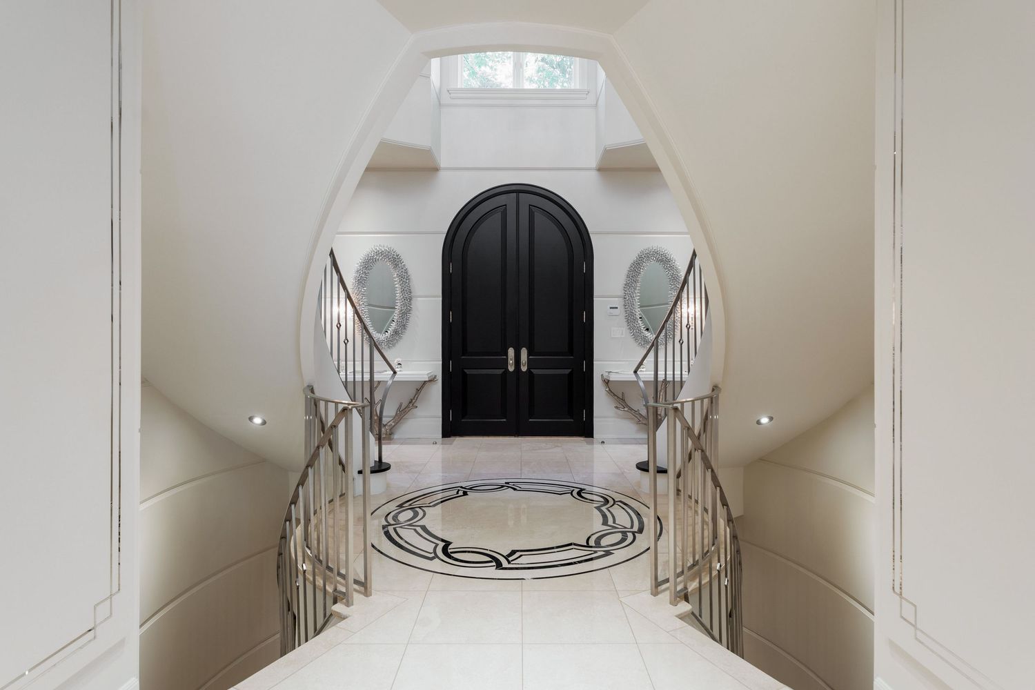 Elegant white marble foyer with curved double staircase and black arched doorway features ornate metal railings.