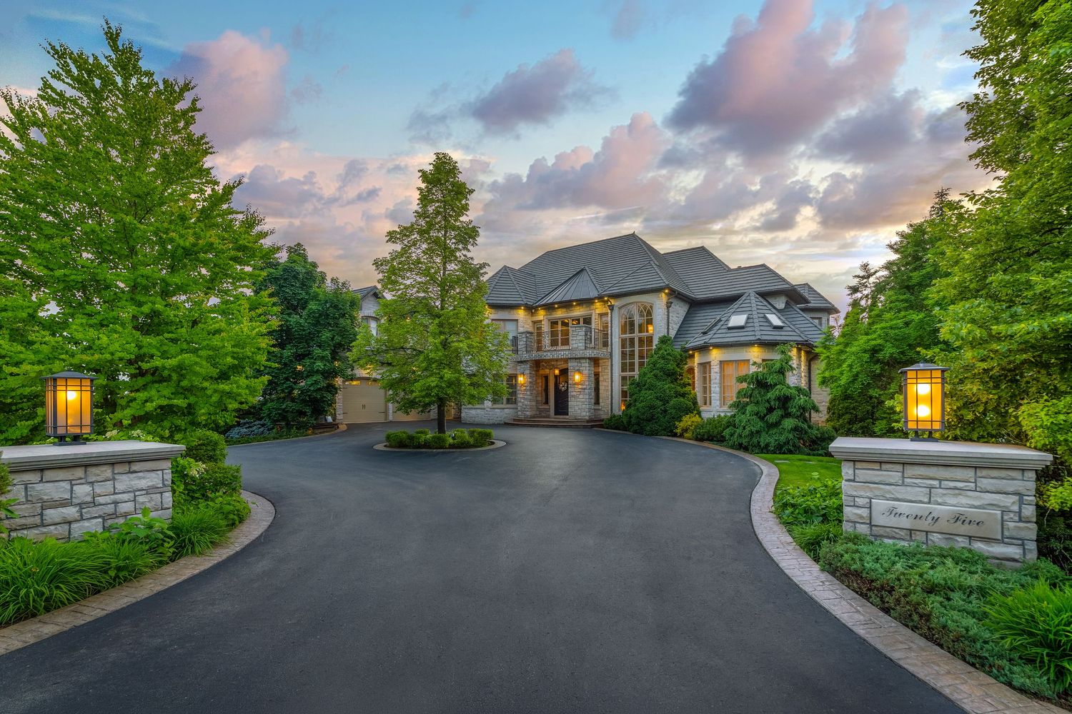 Luxury stone mansion with circular driveway, lush trees, and elegant lantern pillars at dusk.