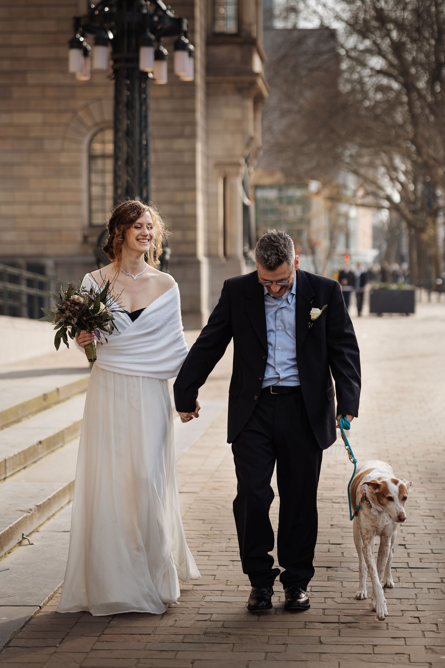 A bride and groom walk with their white dog along a stone pathway.