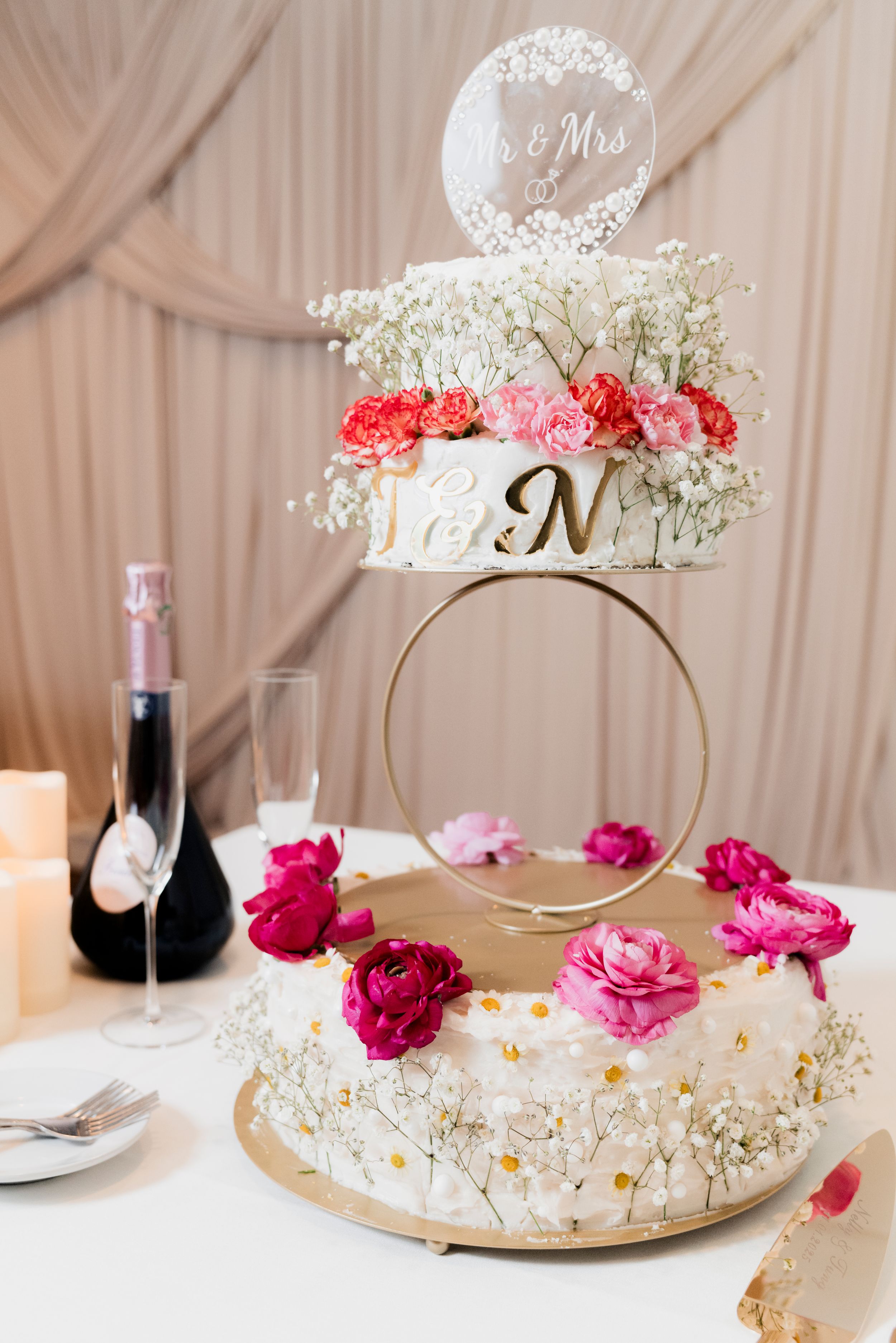 Two-tier white wedding cake adorned with pink roses and baby's breath on a gold ring stand.