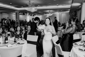 Bride walks through wedding reception hall carrying bouquet while guests celebrate at decorated tables.
