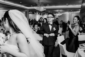 Groom in tuxedo smiles as bride in wedding dress enters elegant reception venue with guests.