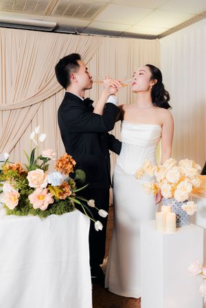 Groom tenderly feeding wedding cake to his bride surrounded by beautiful floral arrangements at reception.