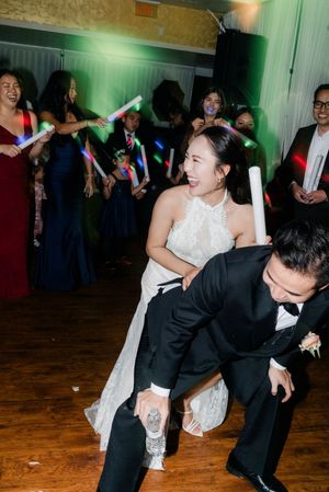 Bride in lace gown and groom in tuxedo dancing with glowing foam sticks at wedding reception.