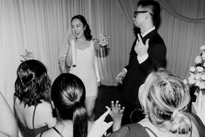 Bride in white dress speaks into microphone while groom gestures to guests at wedding reception.