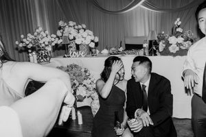 Black and white photo of wedding guests laughing and enjoying drinks at a floral-decorated reception table.