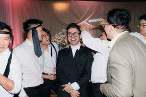 Groom in black suit smiling as groomsmen playfully adjust ties at wedding reception.