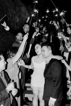 Black and white photo of newlyweds exiting through sparkler arch surrounded by celebrating wedding guests.